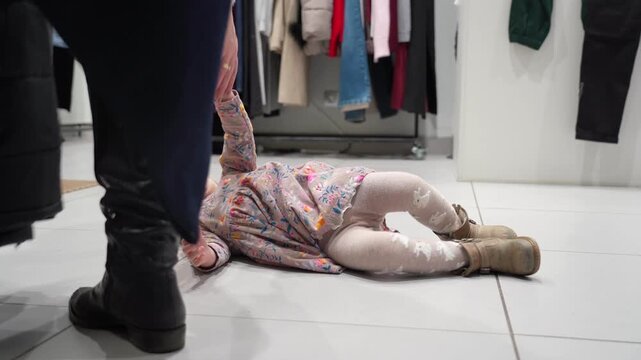Young child in store throws tantrum on floor, upset and crying while lying down amidst clothing racks.