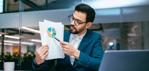 A man in a suit holds documents with charts and graphs while seated at a desk. He focuses on the data as he discusses it with colleagues in a bright office space.