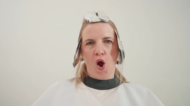 woman at salon with foils, seated in white cape making playful puckered expressions while stylist processes blonde highlights under neutral studio lighting, candid closeups capture reaction,
