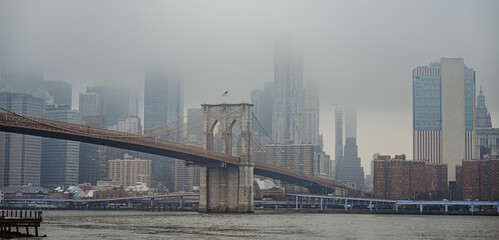 Obraz premium Fog covers Brooklyn Bridge and Manhattan skyline on a cloudy day. Fog fills the air over Brooklyn Bridge and the Manhattan skyline in New York City during an overcast moment.