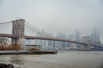 Fototapeta premium Brooklyn Bridge in fog with city skyline and waterfront view. Brooklyn Bridge stands in fog with a view of tall buildings and water.