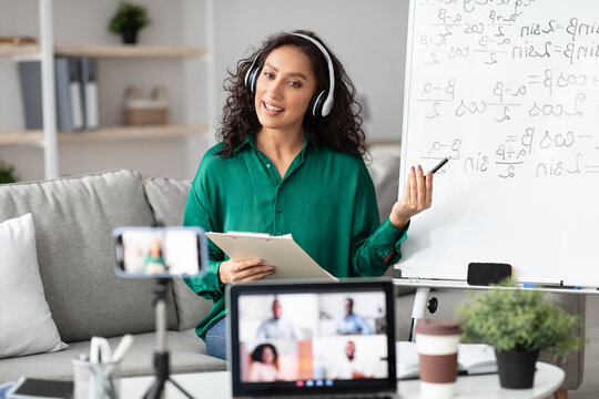 Online Training. Young female tutor in headset teaching online by video conference using laptop and smartphone. Teacher explaining information to diverse participants in the virtual conference - Powered by Adobe