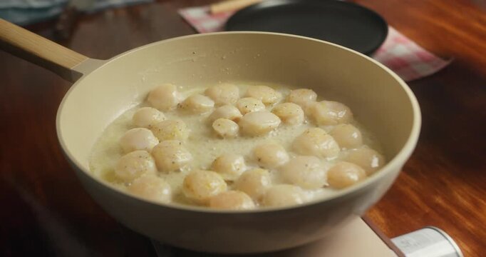 Woman cooking scallops at home in her kitchen, frying sea scallops in a pan on a portable gas stove. Close-up of scallops being turned to achieve an even golden sear, homemade seafood cooking process