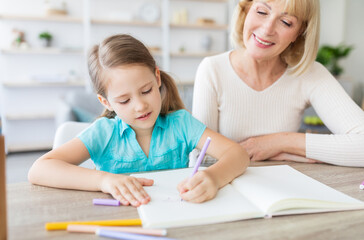 Fototapeta premium Portrait of smilig mature woman helping little girl with homework, spending free time with small preshool grandchild, sitting together at table, drawing pictures using colorful pencils and markers