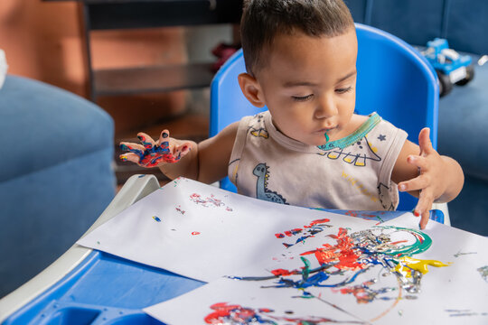 Toddler with paint on hands and mouth playing with colors while sitting indoors at home