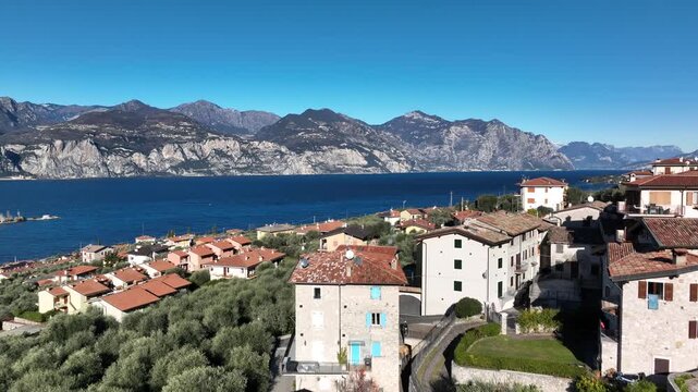 Drone flight over a small Italian village near Brenzone on Lake Garda. Stone houses, bright blue water, mountains and clear sky create a vivid Mediterranean landscape.