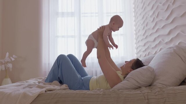 mother lifting baby above chest on bed. morning exercise and play mom with infant with bright window light. cheerful movement and eye contact. happy domestic maternal routine. cozy bonding