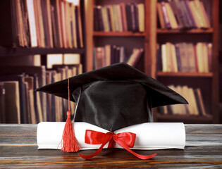 Rolled diploma tied with red ribbon and graduate cap on wooden table in library