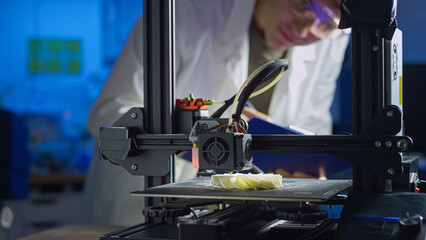 Engineer in lab coat and safety glasses adjusts 3D printer while creating a prototype in a modern lab focused on advanced technology.