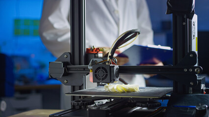 Engineer in lab coat and safety glasses oversees 3D printing process in modern workspace, focusing on precision and technology.