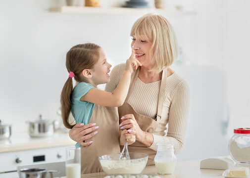 Cooking With Family Is Fun. Portrait of happy mature woman hugging her grandkid baking in kitchen and fooling around, girl playfully touching grandma's nose, kneading dough together and having fun