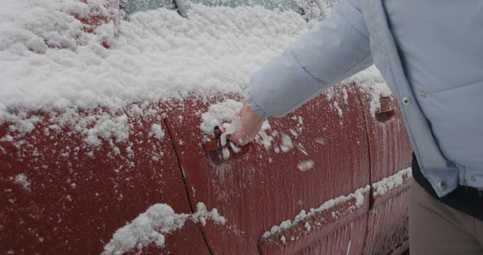 Woman's hand trying to open a red car door handle covered in a thick layer of ice and snow after a winter freezing rain storm, struggling with the frozen lock on a cold day.