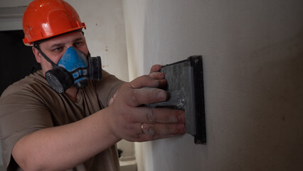 Construction worker sanding a plastered wall surface with a sanding block and mesh abrasive, wearing an orange hard hat and a blue respirator mask for safety during renovation work.