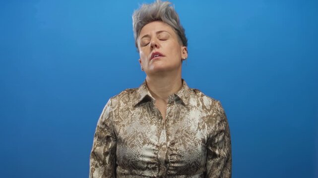 Caucasian woman with short gray hair wearing snake print blouse rolls eyes in solid blue studio; disgust.