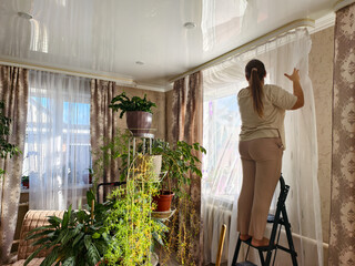 A woman on a stepladder attaches a white translucent curtain to a ceiling cornice above a window. The woman hangs the curtain while standing on the stepladder in a sunlit room. © Александр Лебедько