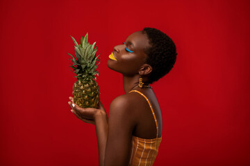 Profile portrait of delighted stylish young african american woman in summer outfit posing with pineapple over red studio background, wearing bright accessories and makeup, copy space © Prostock-studio