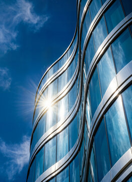 Modern skyscraper glass facade with wavy architectural design reflecting blue sky and bright sun flare creating artistic commercial urban view