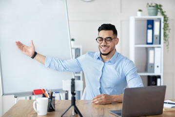 Obraz premium Young Arab male tutor conducting online lesson near empty blackboard, using smartphone for remote education at home office, copy space. Millennial male teacher giving webinar or internet conference