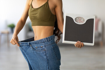 Unrecognizable Indian woman in oversized jeans holding scales, showing results of weight loss program or liposuction at home. Millennial lady achieving success in slimming. Healthy lifestyle concept