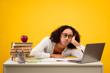 A student rests her head on her hand while sitting at a desk. She is surrounded by books, a laptop,...