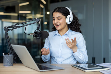 Young woman wearing headphones and speaking into a microphone with animated gestures while recording a podcast or live streaming online from a modern office studio