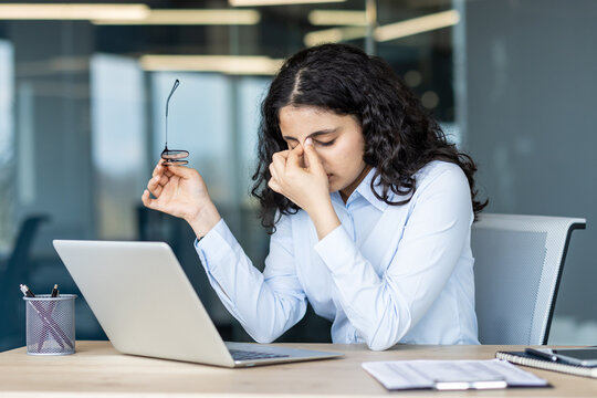Stressed young woman working at office desk and rubbing eyes from headache, feeling tired and burned out, experiencing pressure and eye strain from computer work