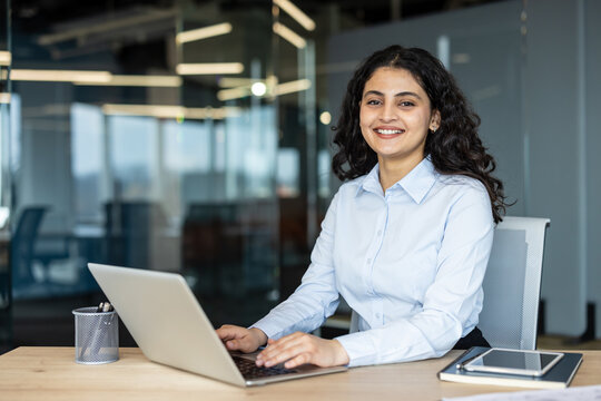 Young indian businesswoman smiles confidently while typing on a laptop at a modern office desk, working on her career and enjoying a productive, professional moment
