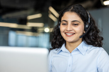 Smiling indian woman wearing a headset and light blue shirt, providing excellent customer service from an office workstation, showing dedication and professionalism