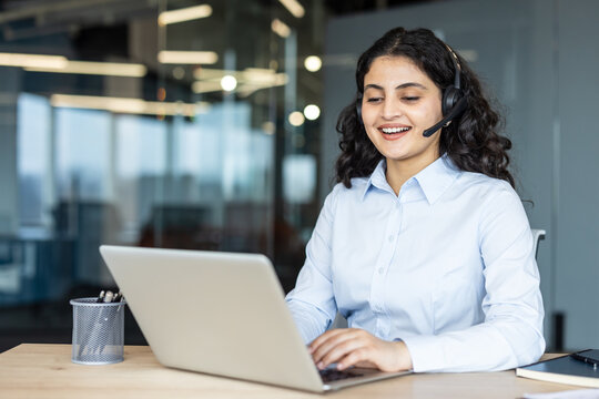 Young indian woman providing customer service and technical support, communicating with clients online through a headset and laptop in a modern call center office