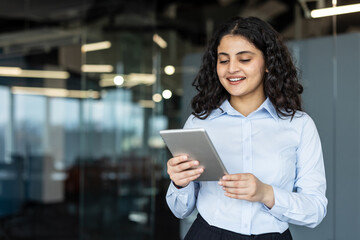 Young professional businesswoman with curly hair smiling while interacting with a digital tablet, standing in a contemporary office reflecting business technology and communication