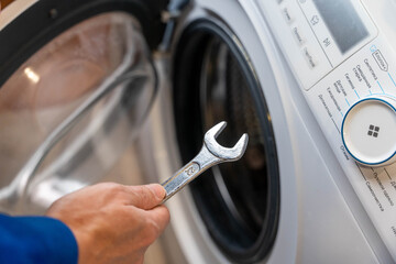 Hand holding a wrench is positioned near the door of a washing machine, showcasing the appliance's control panel and interior during a maintenance task in a home setting