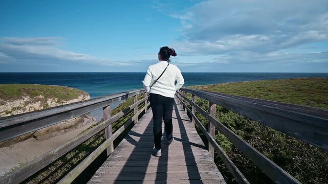 Young woman from behind walking on a long wooden walkway towards the ocean at the famous Las Catedrales beach, in Galicia, Spain, on a sunny day with some clouds and a clear blue sky