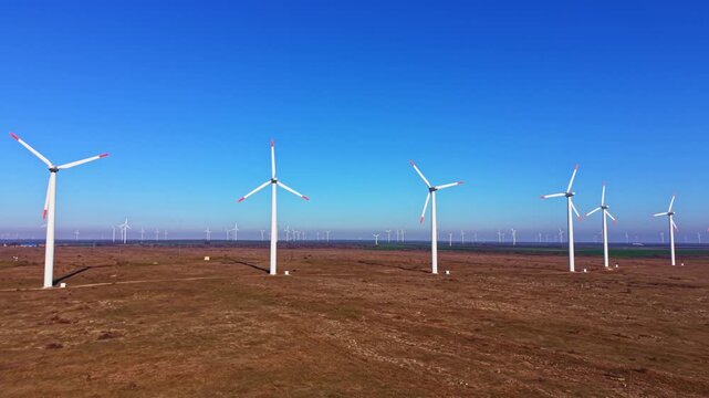 A line of wind turbines is located in an open field under a clear blue sky. Additional turbines can be seen in the distance, creating a large wind farm landscape.