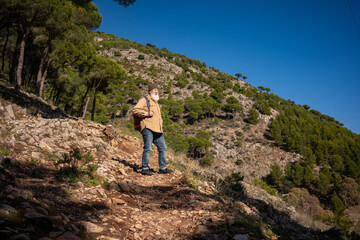Naklejka premium Senior man hiking mountain trail alone, active aging lifestyle, outdoor adventure, healthy retirement, endurance, freedom, independence, walking uphill, forest landscape, sunny day