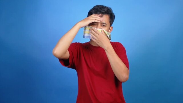 Asian man with flu and headache holding cloth covering nose and other hand holding head isolated on blue background
