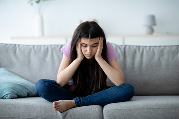 Covid related teen depression concept. Unhappy Indian teenage girl sitting on sofa at home with...