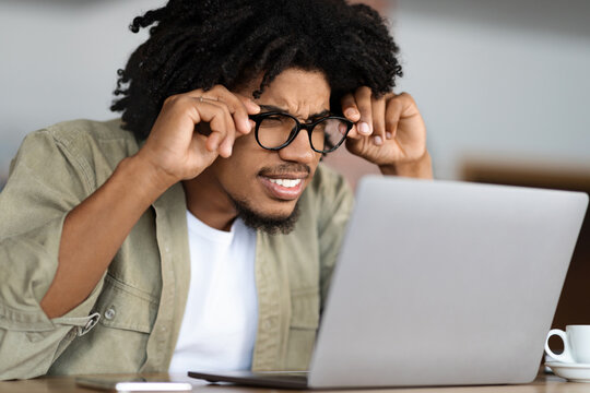 Concentrated young black curly guy in glasses squints and looks at laptop at table with cup in cafe interior. Problems at study and work remotely, independent contractor, overwork and bad eyesight