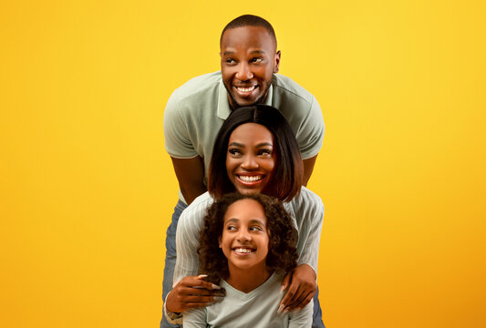 Promo concept. Happy black family of three looking aside at free copy space on yellow studio background, standing behind each other. Empty space for dvertisement