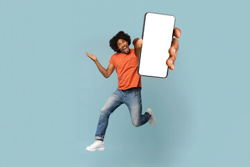 Positive afro-american guy jumping up and showing big smartphone with blank screen, smiling and gesturing, showing newest mobile application, mockup, blue studio background, copy space © Prostock-studio