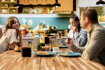 Mother getting distracted by her smartphone and social media addiction at breakfast, being distant with her tween girl and husband. Woman with mobile phone obsession and dependency.