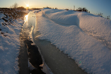 Winter landscape with a frozen stream and melting snow at sunset. Ice, water and sunlight create a natural pattern symbolizing seasonal transition, thaw and environmental change