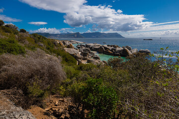Fototapeta premium Penguin Bay near Cape Town shows African penguins waddling along a sandy shore, granite boulders and a wooden boardwalk with visitors, turquoise water, dramatic clouds, and a wild coastal landscape.
