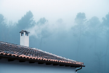 House roof and chimney with smoke rising, surrounded by heavy fog and forest trees creating a cold, mysterious atmosphere