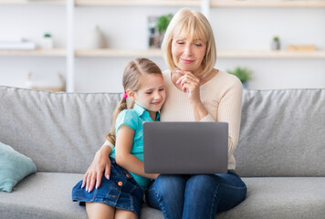 Portrait of smiling senior grandma and cute kid granddaughter watching cartoons on laptop, older grandmother hugging little girl sitting on couch and looking at screen, spending time together