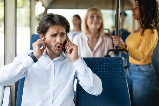 Portrait of confused man talking on the cell phone and holding finger in ear while commuting by public transport, male can't hear because of noise and passengers, bad cellular connection