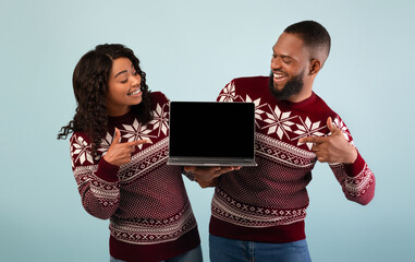 Fototapeta premium Great discounts and sales for New Year. Excited african american couple showing laptop with blank screen, pointing on it and wearing warm sweater on blue background, studio shot