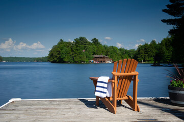 Fototapeta premium A single wooden Muskoka chair on a weathered dock with a white and navy towel, overlooking deep blue lake waters. A cottage is nestled between green trees in the background on a bright afternoon.