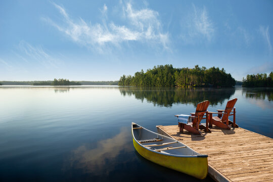 Two wooden Adirondack chairs on a weathered dock in Muskoka, ON, with a blue towel. A yellow canoe rests on the mirror-like lake, with green islands and a clear blue sky reflecting calm cottage life