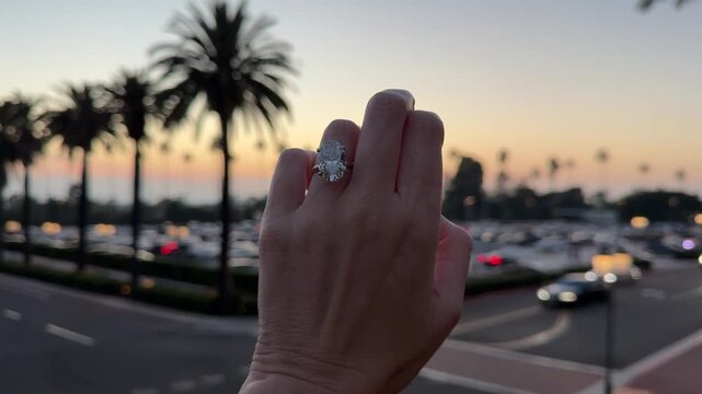 Large oval diamond solitaire engagement ring displayed on a hand with palm trees and sunset sky in Newport Beach.
