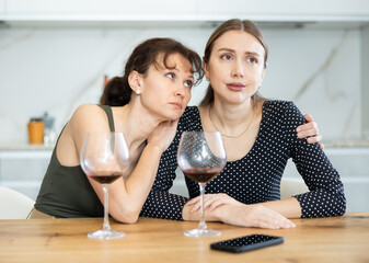 Upset woman in her middle years is seated at table with a wineglass accompanied by another woman offering her support